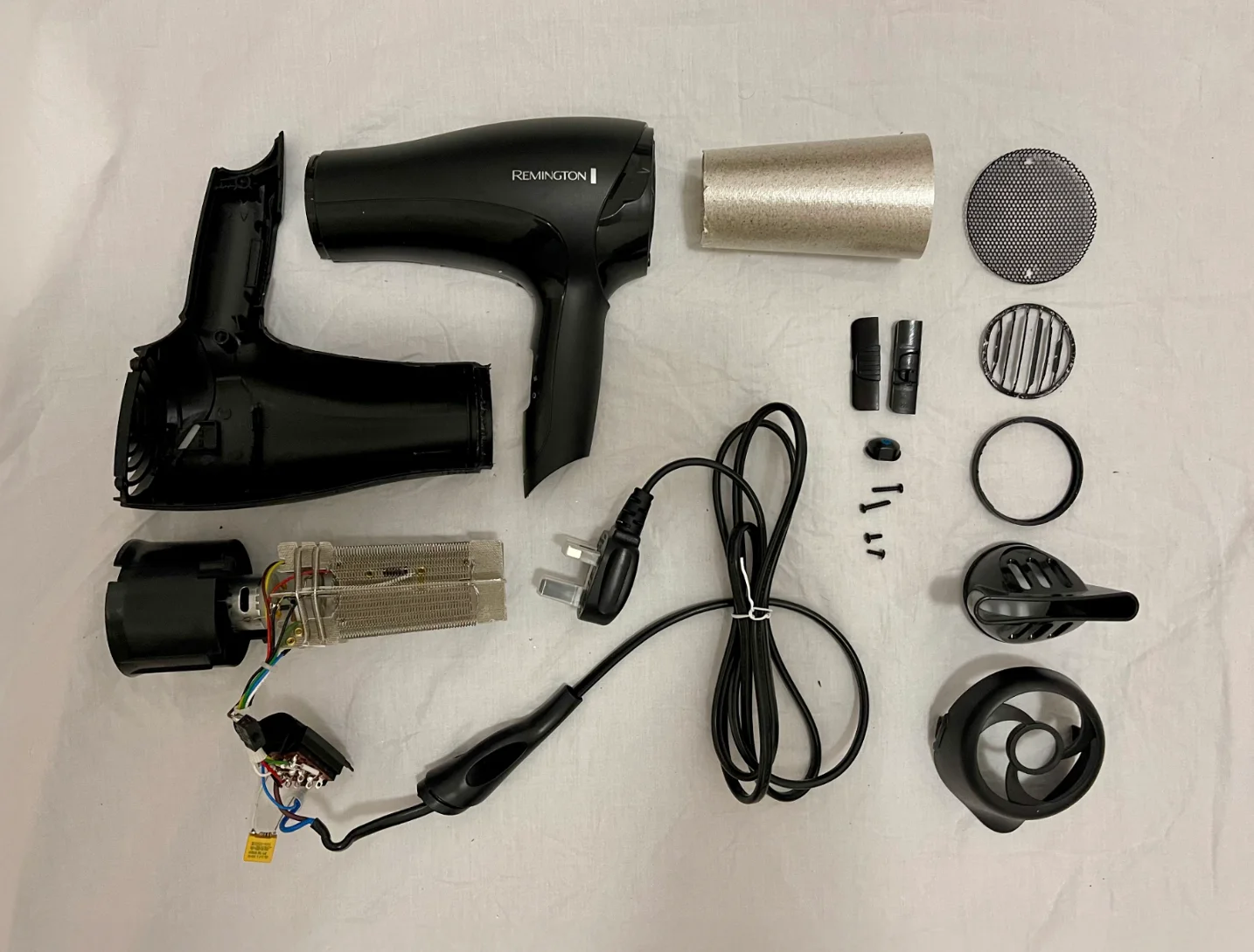 A photograph of the Remington hair dryer fully disassembled, with each component laid out: motor, fan, heating coil, plastic case halves, wiring, screws and grills