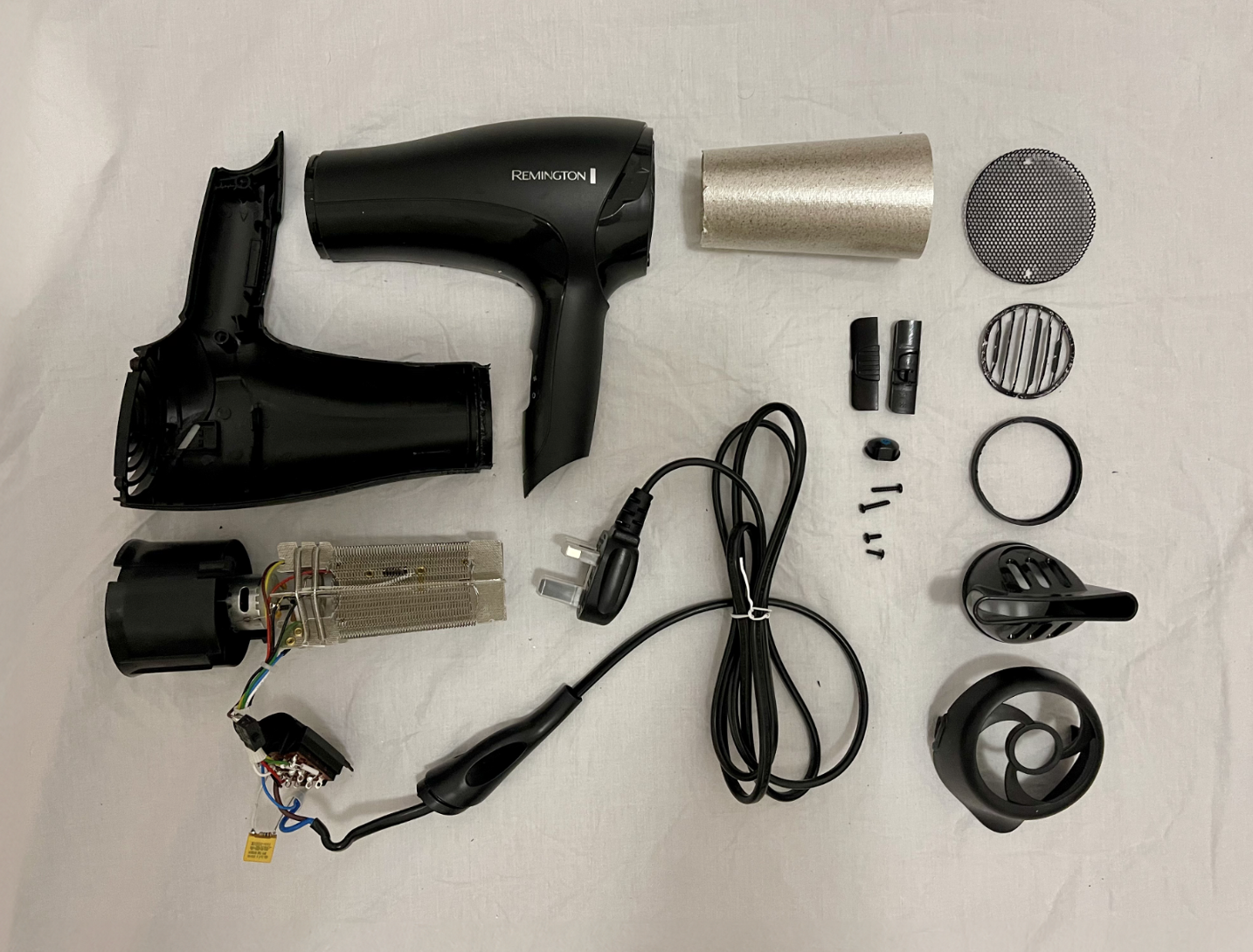 A photograph of the Remington hair dryer fully disassembled, with each component laid out: motor, fan, heating coil, plastic case halves, wiring, screws and grills