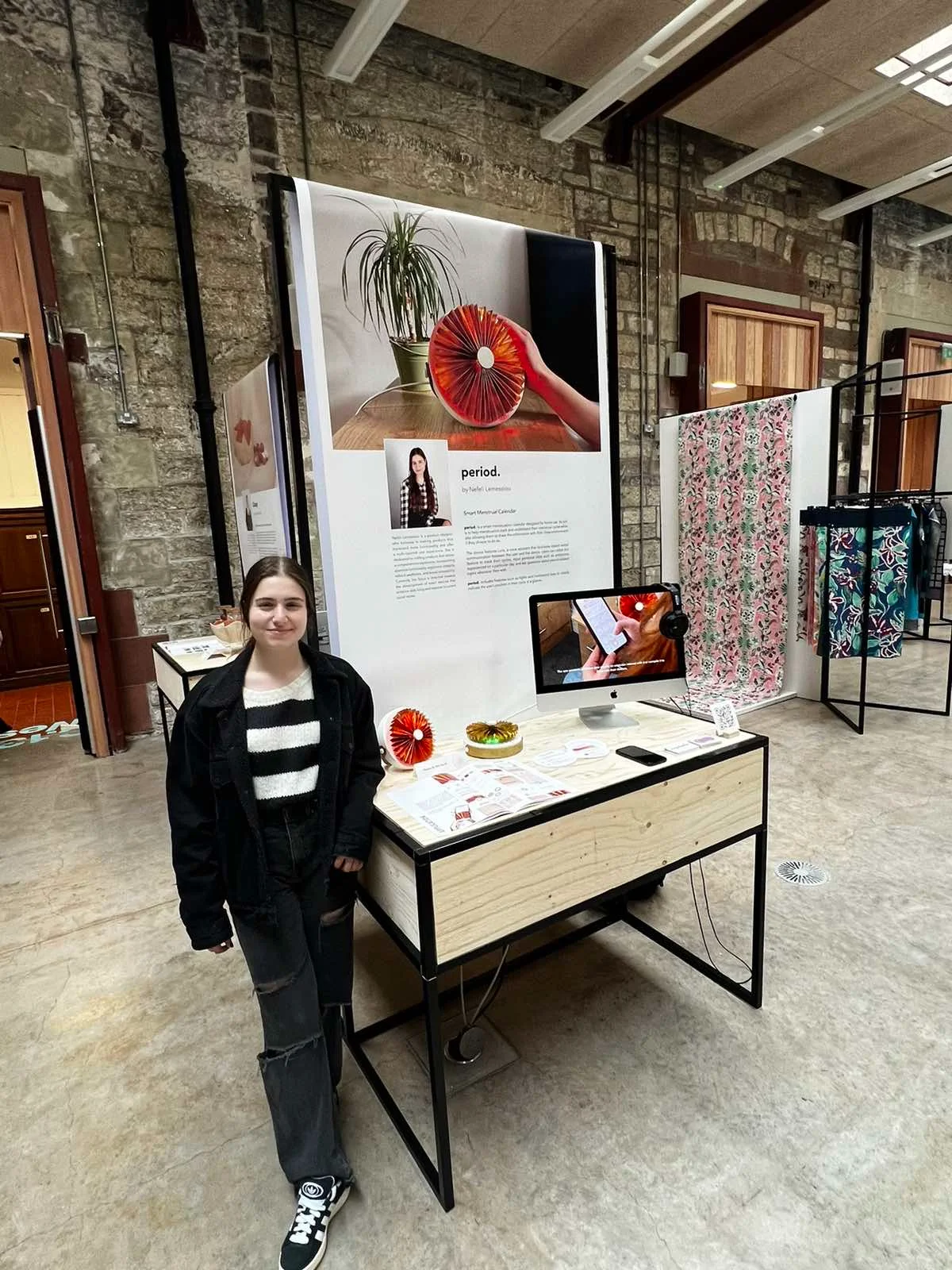 Nefeli standing beside her period. stand at the ECA Graduate Show, with the device, booklet and app on a plywood plinth under a large period. poster