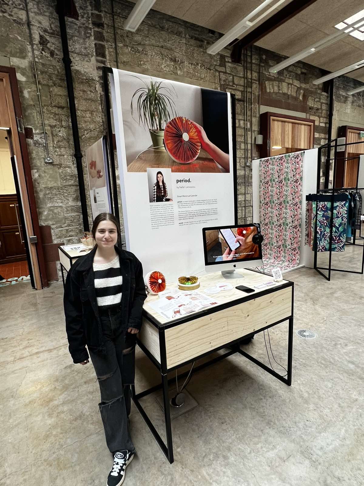 Nefeli standing beside her period. stand at the ECA Graduate Show, with the device, booklet and app on a plywood plinth under a large period. poster