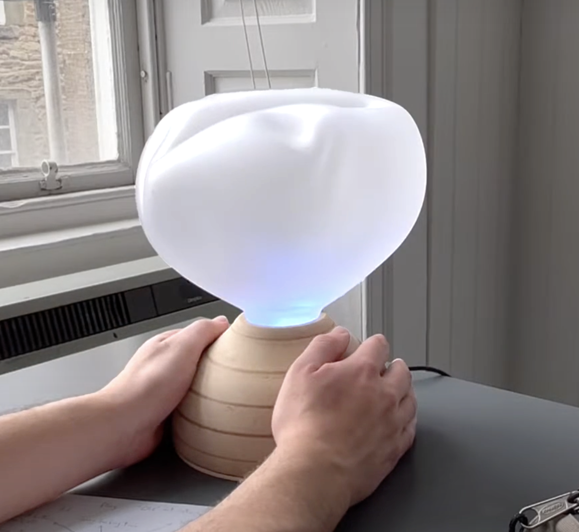 Ilia on a study desk, a sandblasted glass cloud diffuser on a wooden dome base, the diffuser glowing softly with the user's hands placed on either side of the base