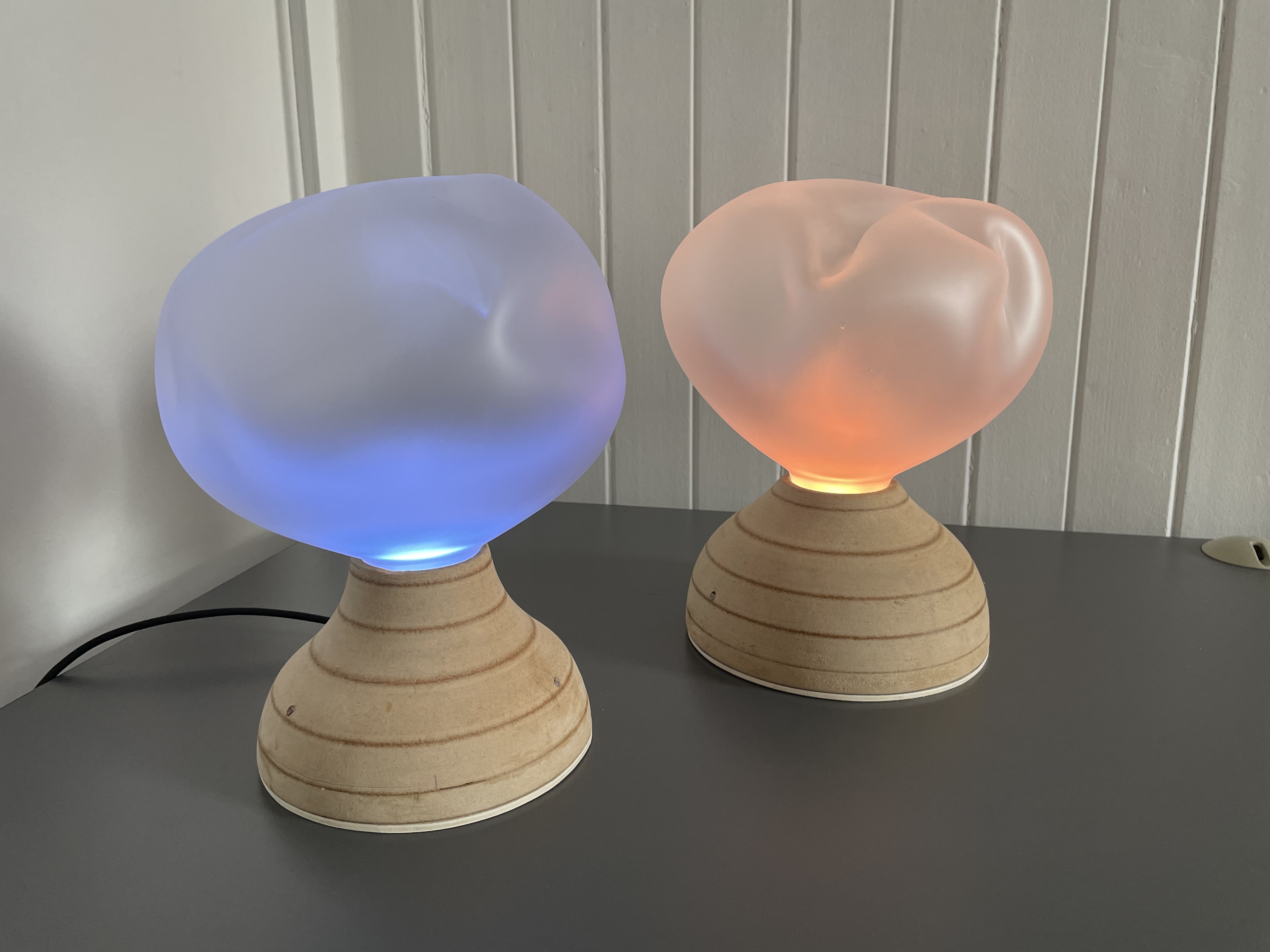 The Ilia pair on a desk — two turned-wood bases with sandblasted glass cloud diffusers, one glowing blue and one glowing warm orange
