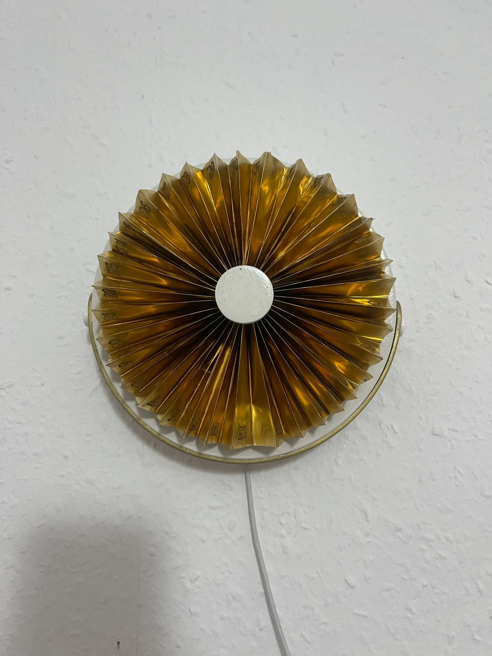 The brass device wall-mounted on a textured white wall, viewed straight on, with the power cable dropping below