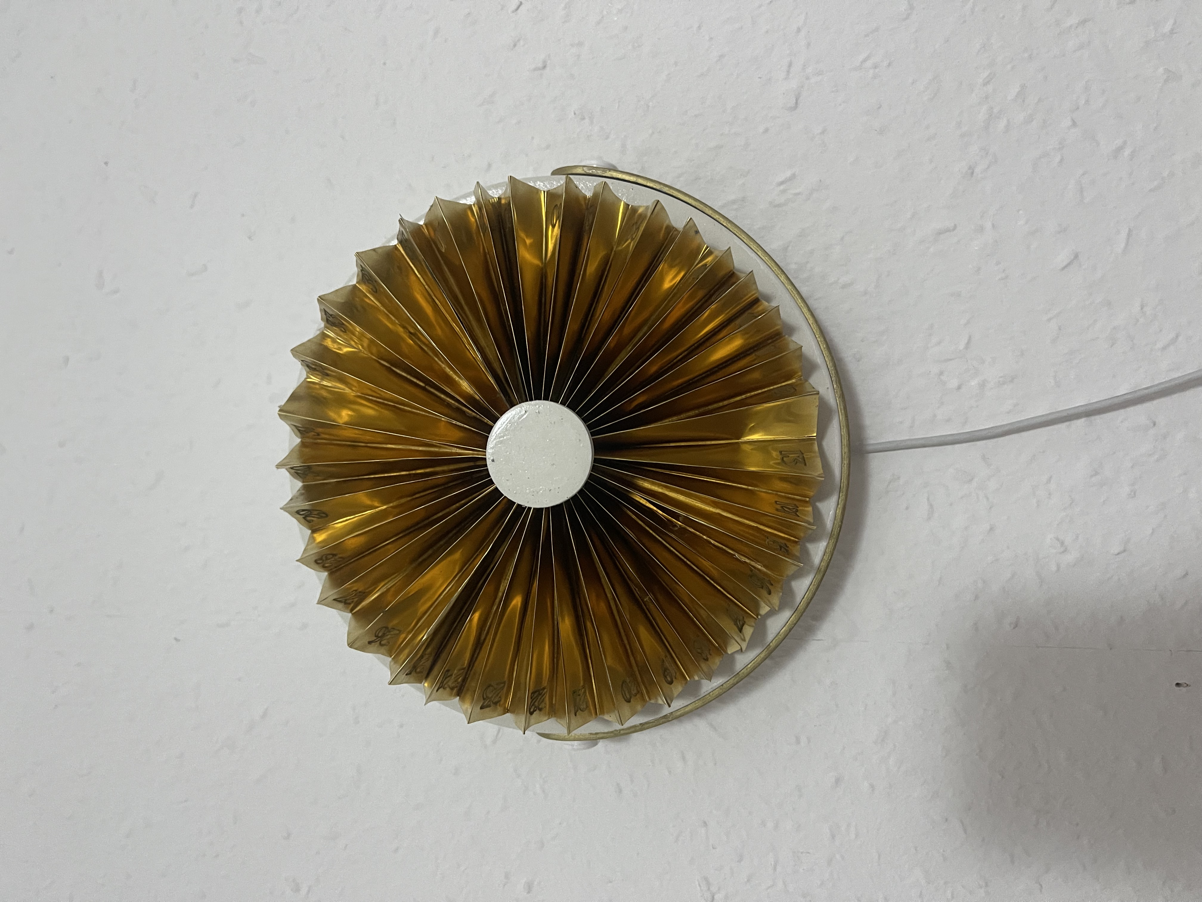 The brass device wall-mounted on a textured white wall, viewed straight on, with the power cable dropping below
