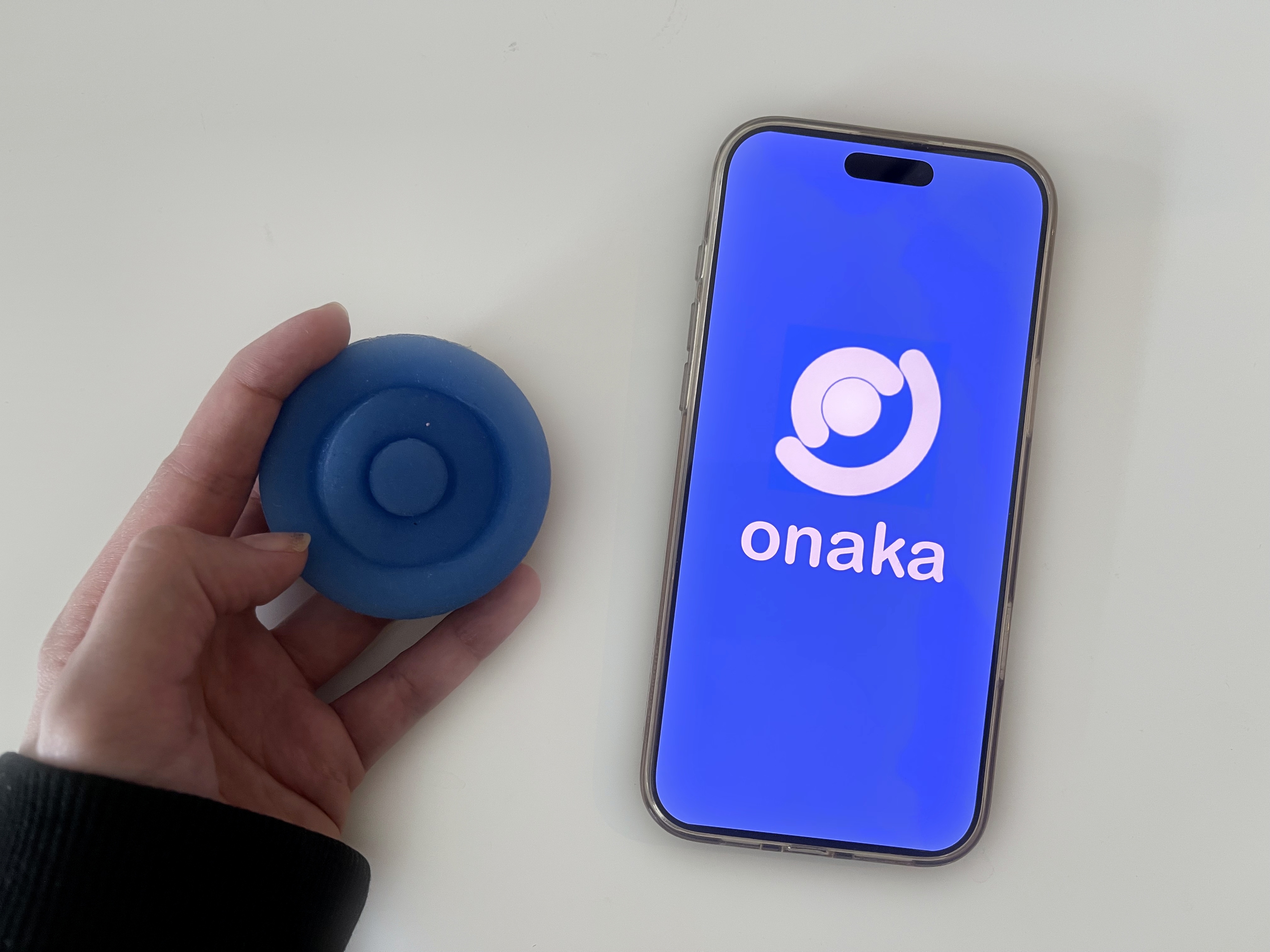 A hand holding the blue Onaka device beside a phone showing the onaka app splash screen